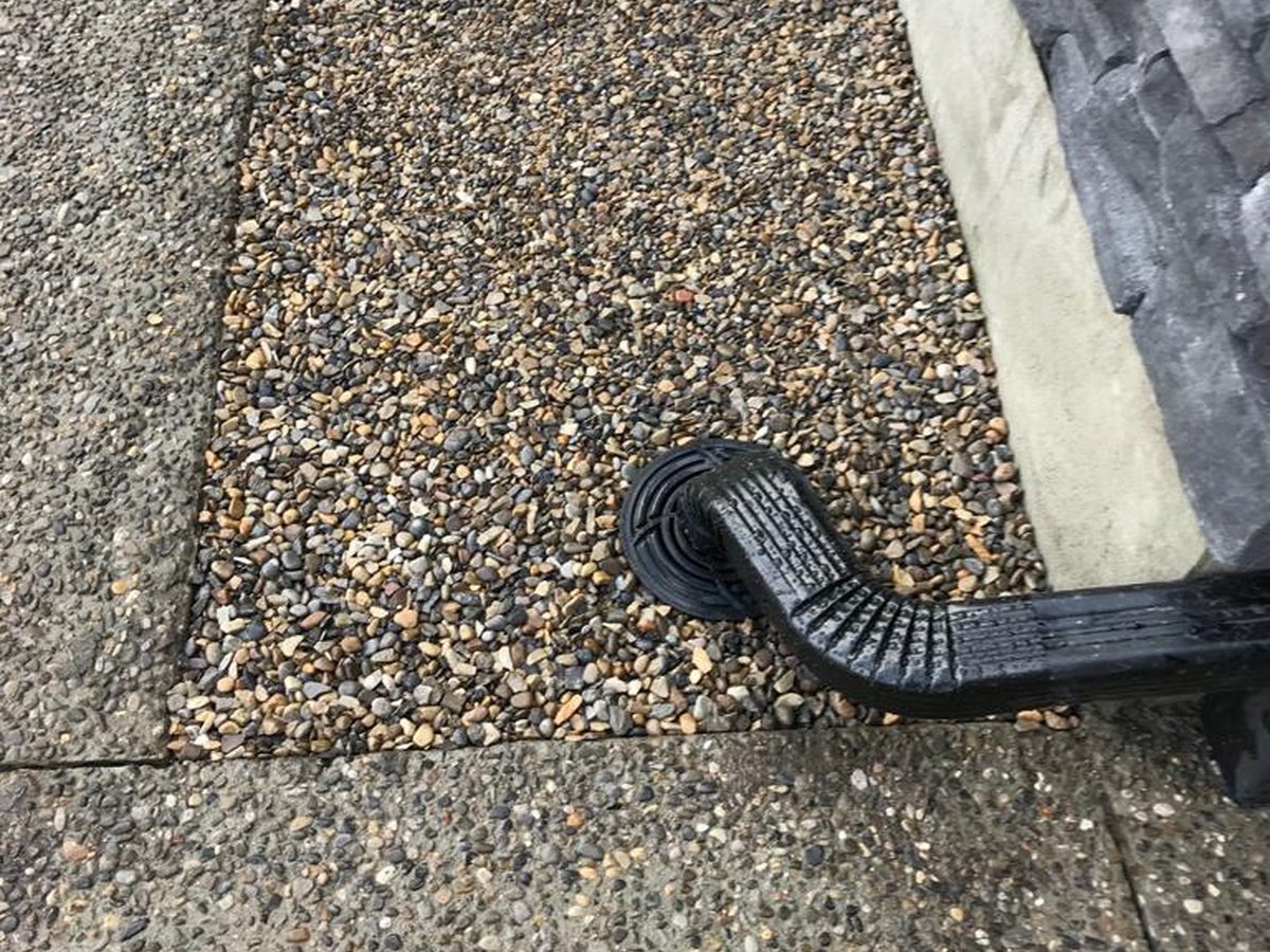 Black downspout extension carrying roof water across gravel away from a house foundation